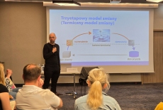 Uczestnicy siedzą na krzesłach i dyskutują. Prowadzący stoi.