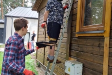 a zielonej trawie stoi brązowy, drewniany dom, a przy nim na drabinie stoi mężczyzna, drugi podaje mu farbę.