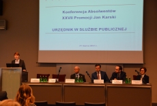 Pani stoi przy mównicy i przemawia, obok przy stole prezydialnym siedzą prelegenci konferencji