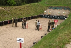 Czterech słuchaczy, z asystą żołnierzy, stoi na strzelnicy i oddają strzały.