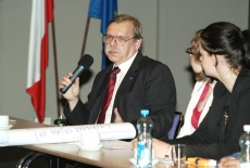 Jeden z uczestników siedzi w ławce i i mówi do mikrofonu, w tle flagi Polski i UE