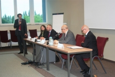 Cztery osoby siedzą przy stole prezydialnym, a jedna osoba przemawia do mikrofonu stojąc. 