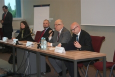 Cztery osoby siedzą przy stole prezydialnym, a jedna osoba przemawia do mikrofonu stojąc. 