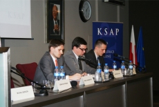 Trzej uczestnicy konferencji siedzący za stołem prezydialnym.