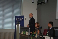 Pan stoi na mównicy i przemawia obok siedzą prowadzący  konferecję