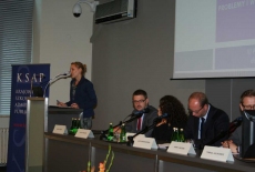 Pani stoi na mównicy i przemawia obok siedzą prowadzący konferecję