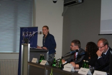 Pani stoi na mównicy i przemawia obok siedzą prowadzący konferecję