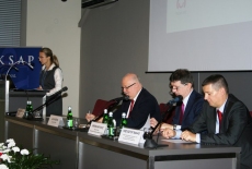 Prelegenci siedzą przy stole prezydialnym. Na mówinicy stoi Elżbieta Kowalewska, pracownik KSAP.