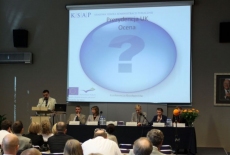 Pan stoi na mównicy i przemawia  za nim wyświetlana jest prezentacja