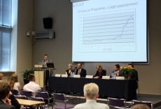 Pan stoi na mównicy i przemawia  za nim wyświetlana jest prezentacja