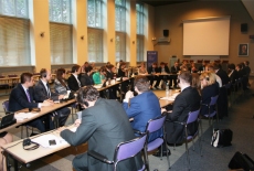 Widok na salę w której odbywa się konferencja. Uczestnicy siedzą przy stołach ustawionych w trójkąt.