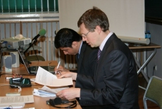 Dwóch prowadzących siedzi przy stole jeden z nich trzyma dokumenty drugi notuje