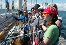 Siedmioro uczestników rejsu stoi w rzędzie wzdłuż rei. Żagle są opuszczone.