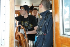 Trzech uczestników rejsu na mostku kapitańskim. Jeden z mężczyzn trzyma koło sterowe.