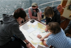 Pięcioro uczestników rejsu pochylonych nad mapą. Jedna z osób wytycza trasę rejsu.