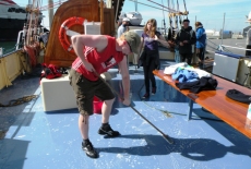 Uczestnik rejsu czyści pokład żaglowca. Statek zacumowany w porcie widać inne jednostki i część nadbrzeża.