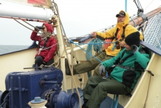 Troje uczestników w kurtkach siedzi na dziobie żaglowca