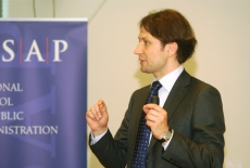 Uczestnik wizyty studyjnej żywo gestykuluje stojąc obok plakatu promującego szkołe i wydarzenie.