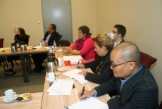 Uczestnicy programu siedzą przy stole ustawionym w podkowę. Słuchają i notują.