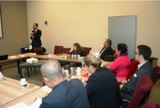 Uczestnicy programu siedzą przy stole ustawionym w podkowę. Jedna osoba stojąc mówi do mikrofonu.