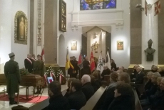 Trumna wystawiona w kościele podszas mszy pogrzebowej. W kościele zgromadzili się ludzie. Stoją poczty sztandarowe.