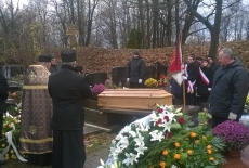 Trumna wkładana jest do grobu. W koło niej zgromadzeni są ludzie i ksiądz.