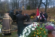 Trumna wkładana jest do grobu. W koło niej zgromadzeni są ludzie i ksiądz.