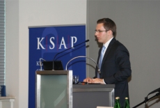 Jeden z uczestników uroczystości przemawia do zgromadzonych z podium. Za nim plakat promujący krajową szkołę administracji publicznej.