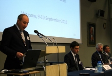 Pan stoi przy mównicy i przemawia obok przy stole siedzą prowadzący seminarium