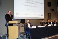 Pan stoi przy mównicy i przemawia obok przy stole siedzą prowadzący konferencje