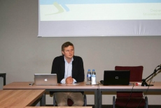 Pan siedzi w ławce i przemawia  w tle prezentacja