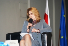 Prelegentka siedząc w fotelu mówi do mikrofonu. Za nią flaga Polski i Uni Europejskiej.