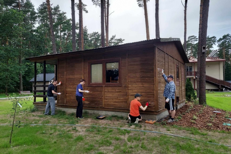 Na zielonej trawie stoi brązowy, drewniany dom, a przy nim czterech mężczyzn maluje ściany