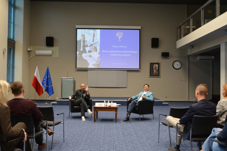 na wprost siedzą na fotelach prowadzący. Tyłem siedzą ludzie zgromadzeni w auli.